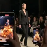 Seorang model berjalan di catwalk mengenakan setelan hitam polos yang berubah menjadi gaun api menyala saat dilihat melalui layar ponsel.